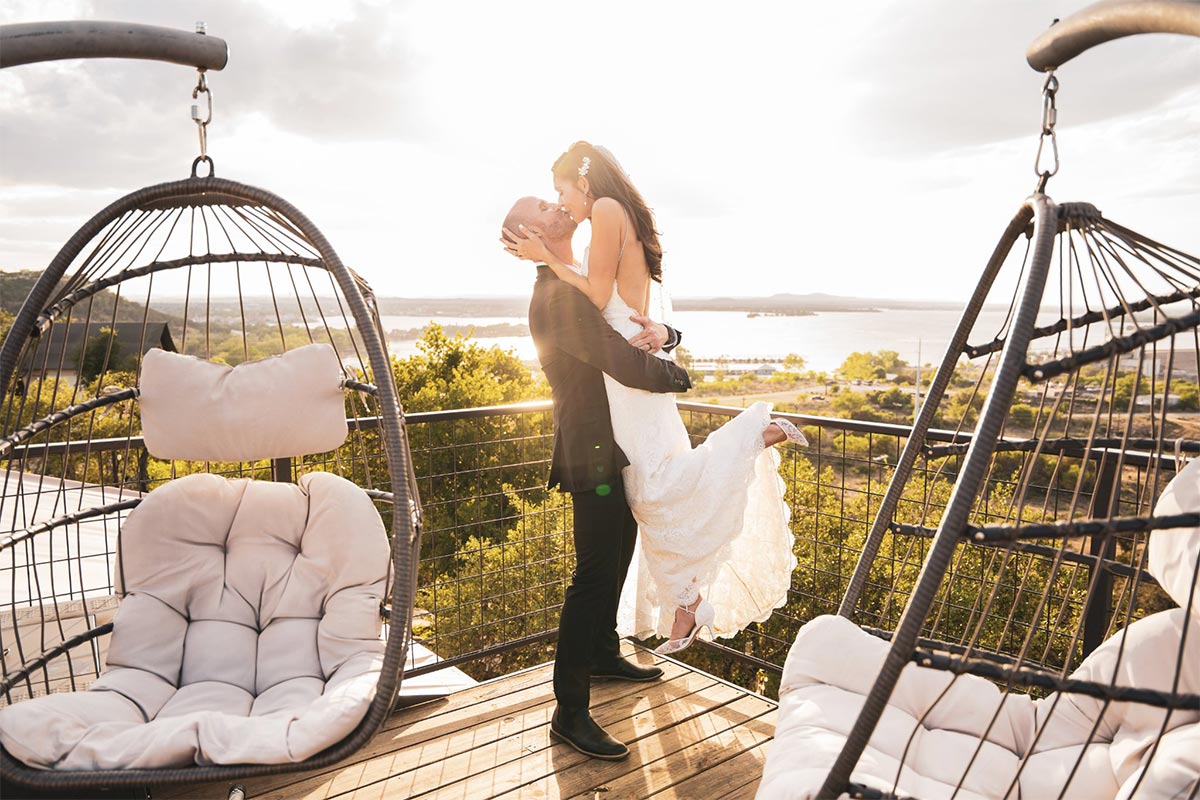 A couple kissing at sunset between hanging egg chairs on a deck above the lake